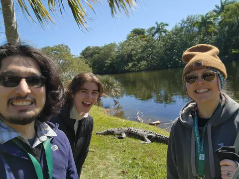 Three lab members take a selfie on a pond bank at Fairchild Tropical Botanic Garden, with a crocodile basking on the grass behind them